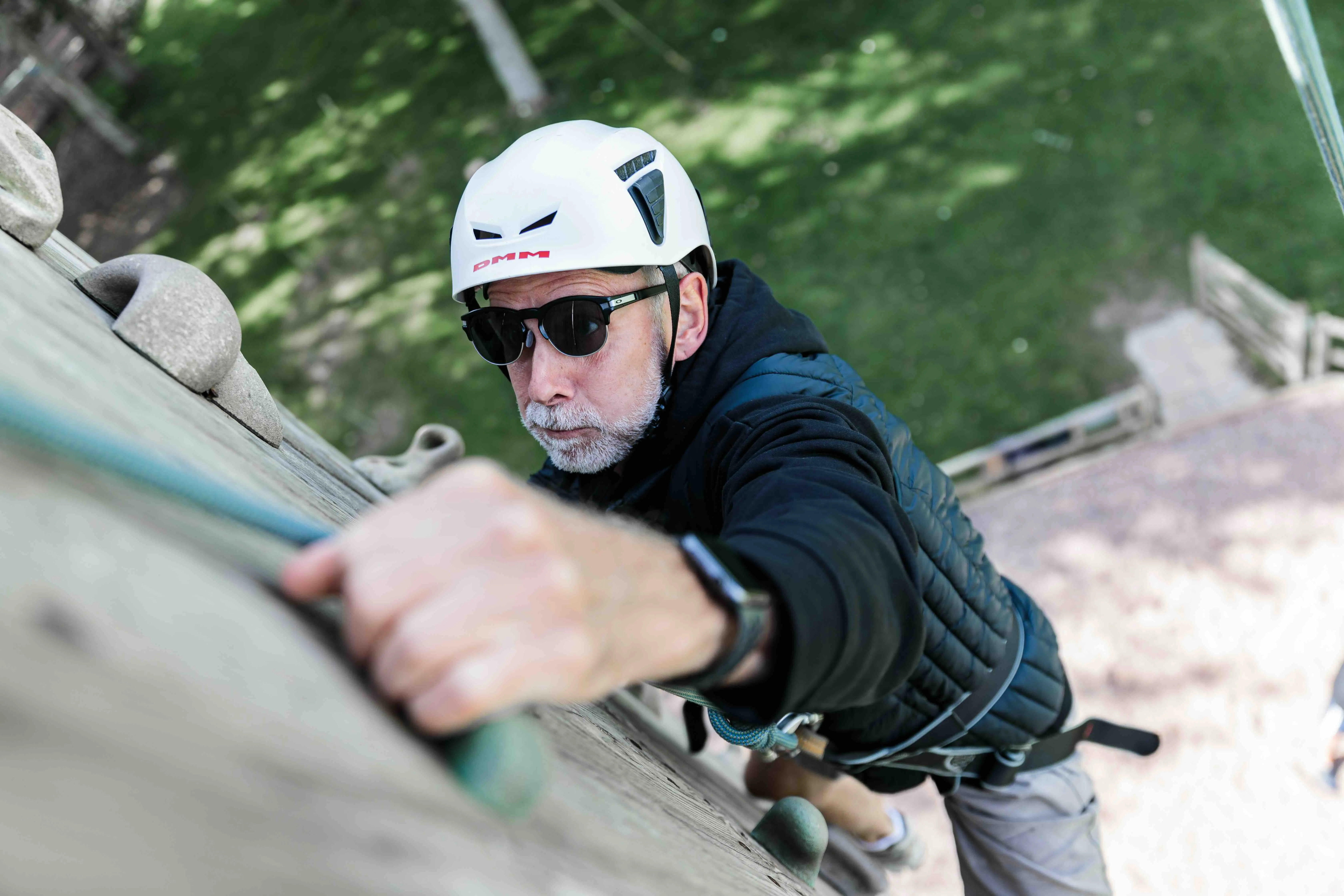 Middle-aged man wearing a white helmet and sunglasses climbing a wooden rock wall outdoors.