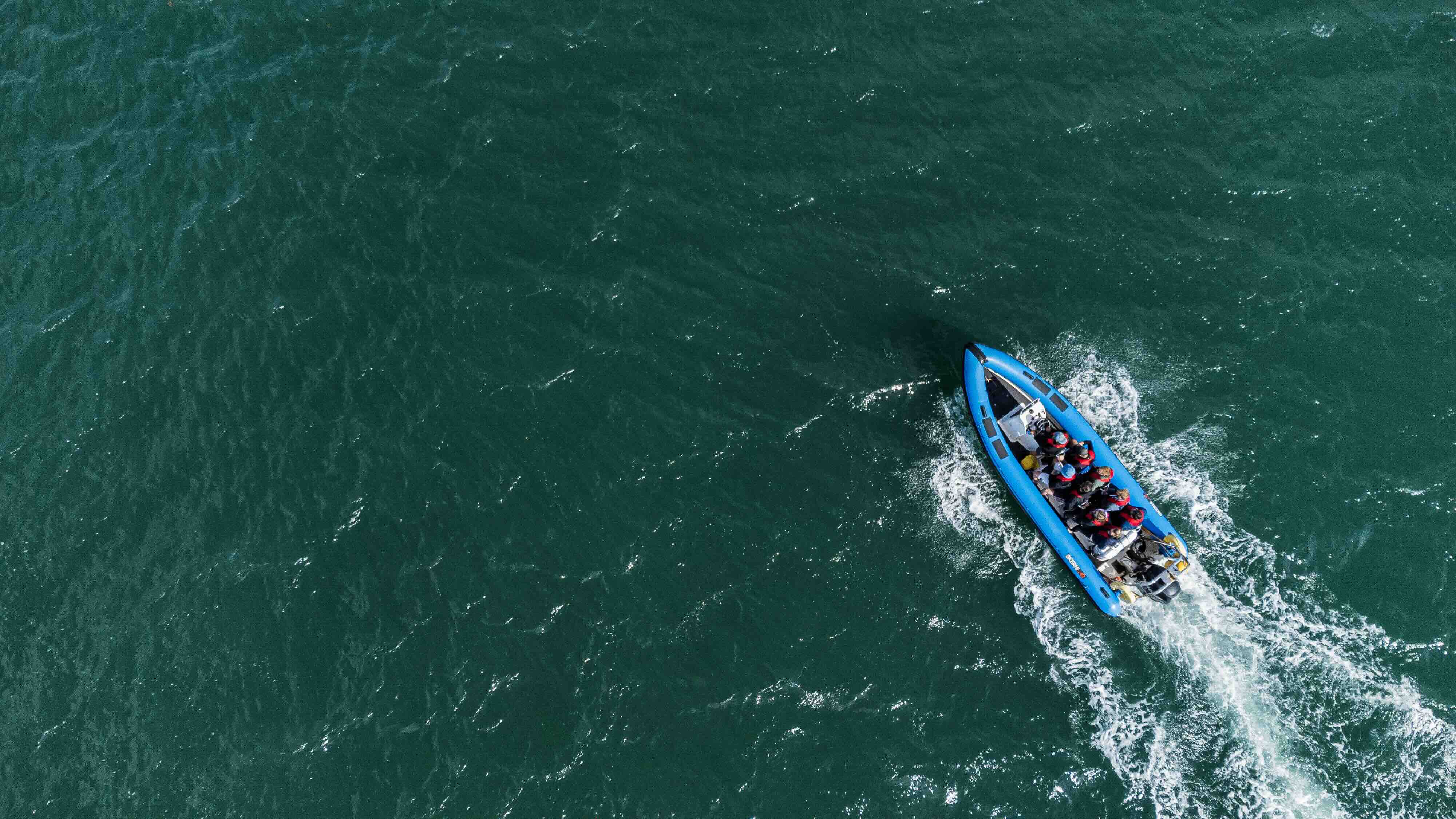 Aerial view of a group of people riding a blue motorized inflatable boat on dark green water.