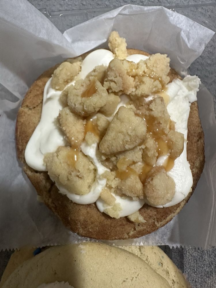 Close-up of a cookie topped with white frosting, crumbly streusel pieces, and caramel drizzle, on parchment paper.