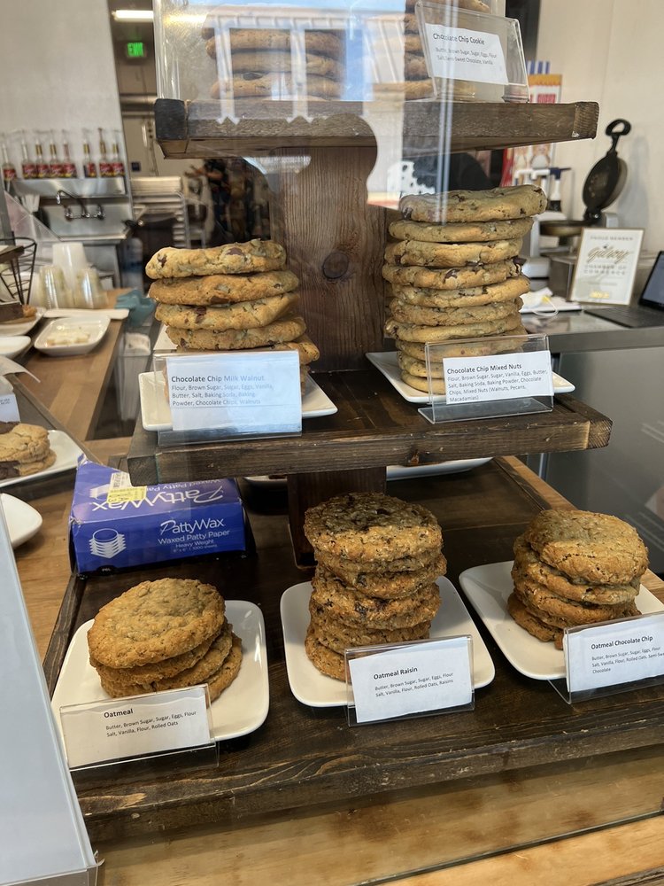 Wooden display shelf with plates of stacked cookies in flavors including Chocolate Chip Milk Walnut, Chocolate Chip Mixed Nuts, Oatmeal, Oatmeal Raisin, and Oatmeal Chocolate Chip, each labeled with ingredient cards.