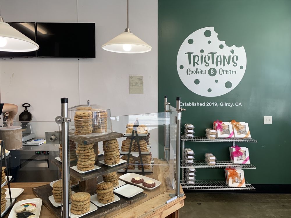 Cookie display case with stacks of cookies on white plates inside a bakery named Tristan's Cookies & Cream, with packaged cookies on shelves against a green wall.
