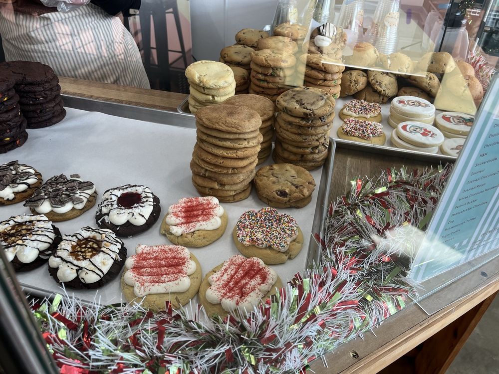 Display case with various stacked and decorated cookies including chocolate chip, frosted with sprinkles, and cookies topped with red sprinkles and white icing.
