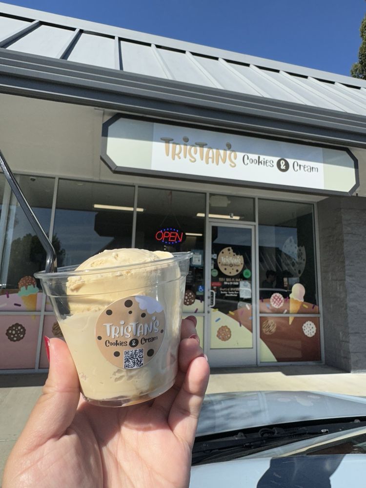 Hand holding a cup of light-colored ice cream with a spoon in front of Tristan's Cookies & Cream shop.