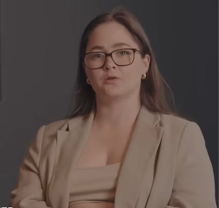 Woman with glasses and brown hair wearing beige blazer and top against a dark background.