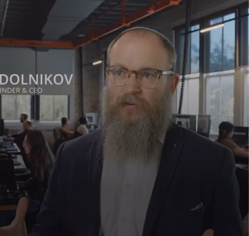 Bearded man with glasses speaking in a modern office with people working at computers.