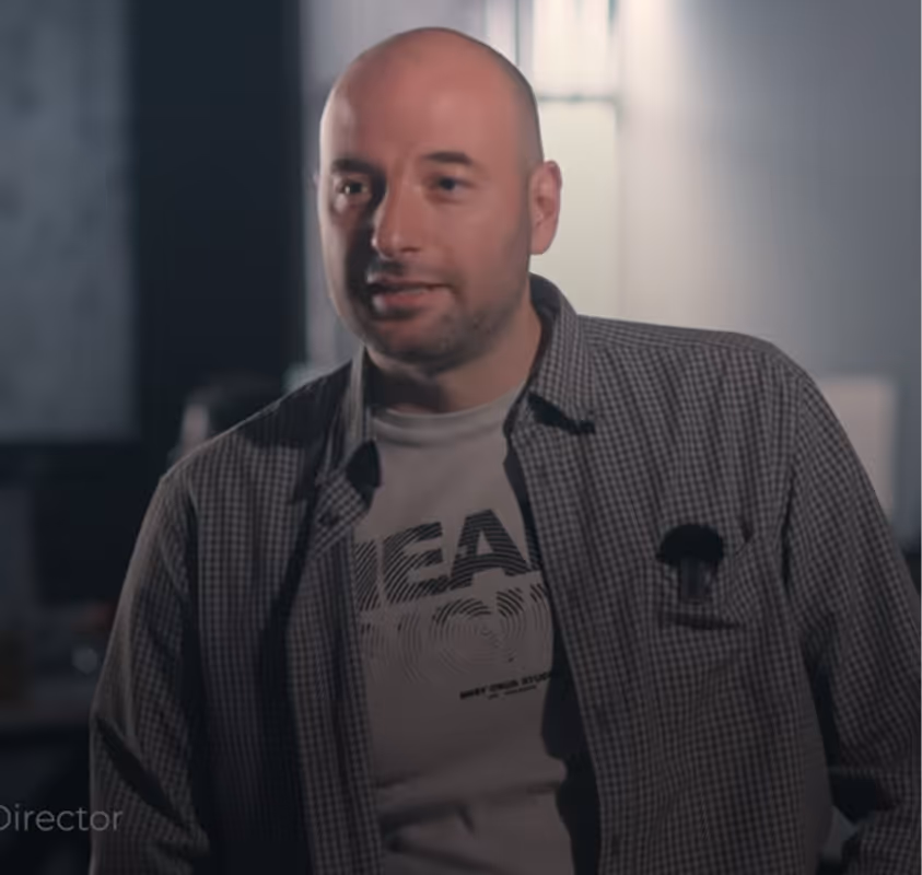 Bald man wearing a checkered shirt over a graphic tee in a dimly lit room.