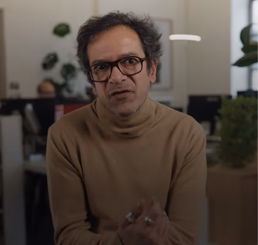 Man with glasses and curly hair wearing a brown turtleneck speaking indoors with blurred plants behind him.