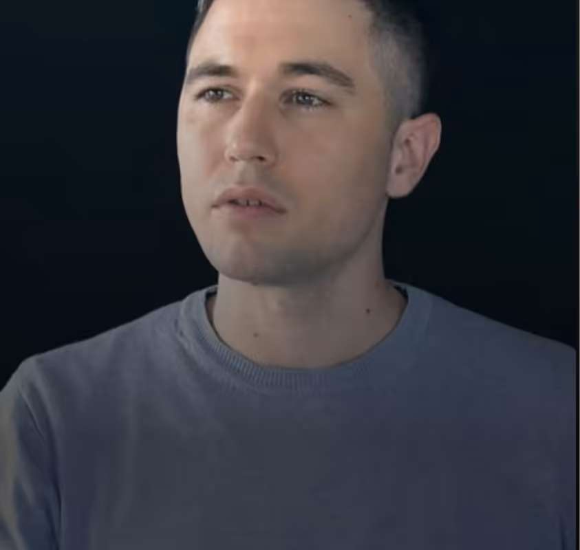 Portrait of a young man with short dark hair wearing a gray sweatshirt, against a black background.