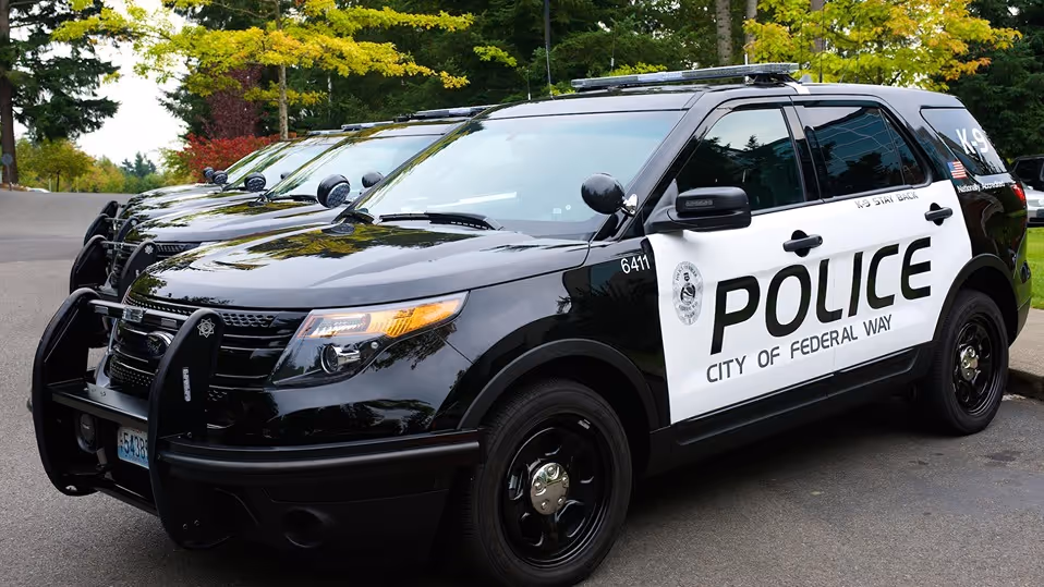 Black and white police SUVs from the City of Federal Way parked in a row on a street with green trees in the background.