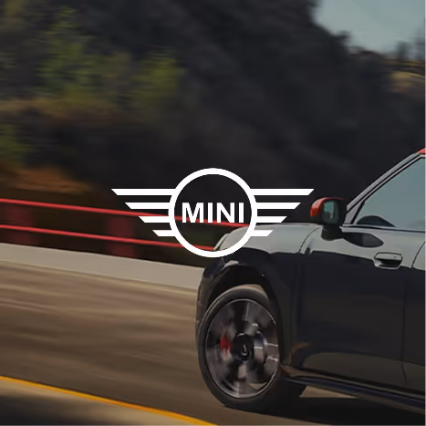 Close-up of a black MINI car driving on a road with a blurred natural background and the MINI logo centered.