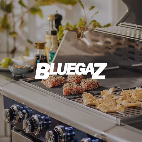 Gas grill with food cooking on the grates, including seasoned meat and sliced mushrooms, with bottles and plants in the background.