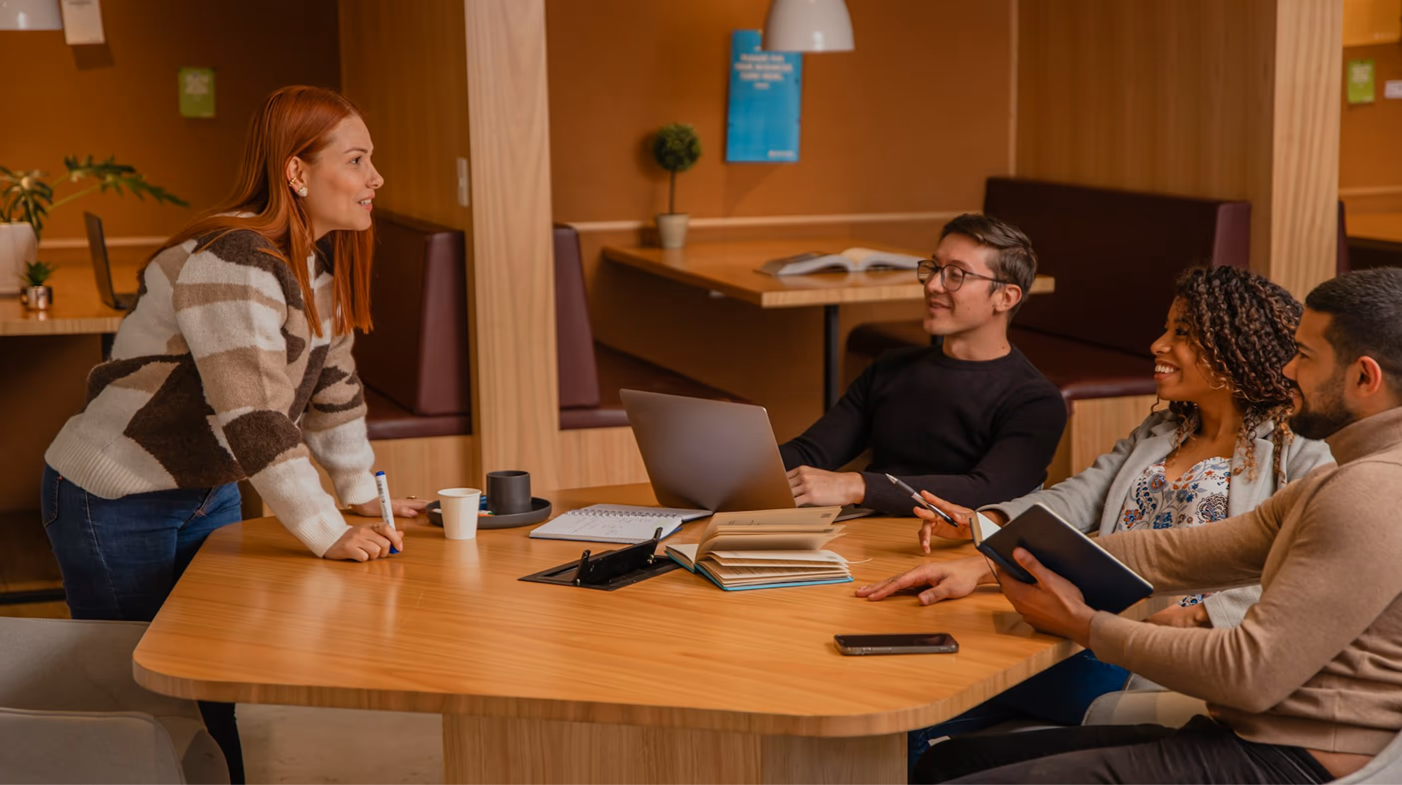 Four colleagues having a discussion around a wooden table with laptops, notebooks, and coffee cups in a casual office setting.