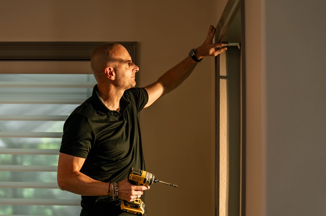 Installer holding a mounting bracket at the top of a window frame with a cordless drill in hand