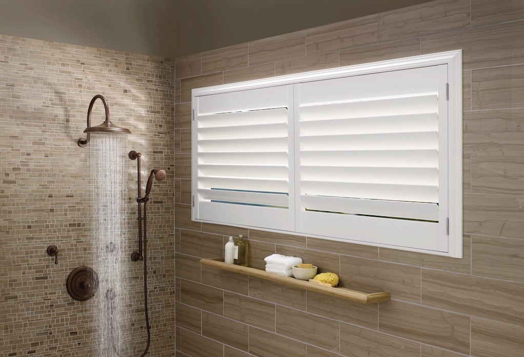 White composite shutters with closed louvers on a tiled bathroom window beside a running rain shower