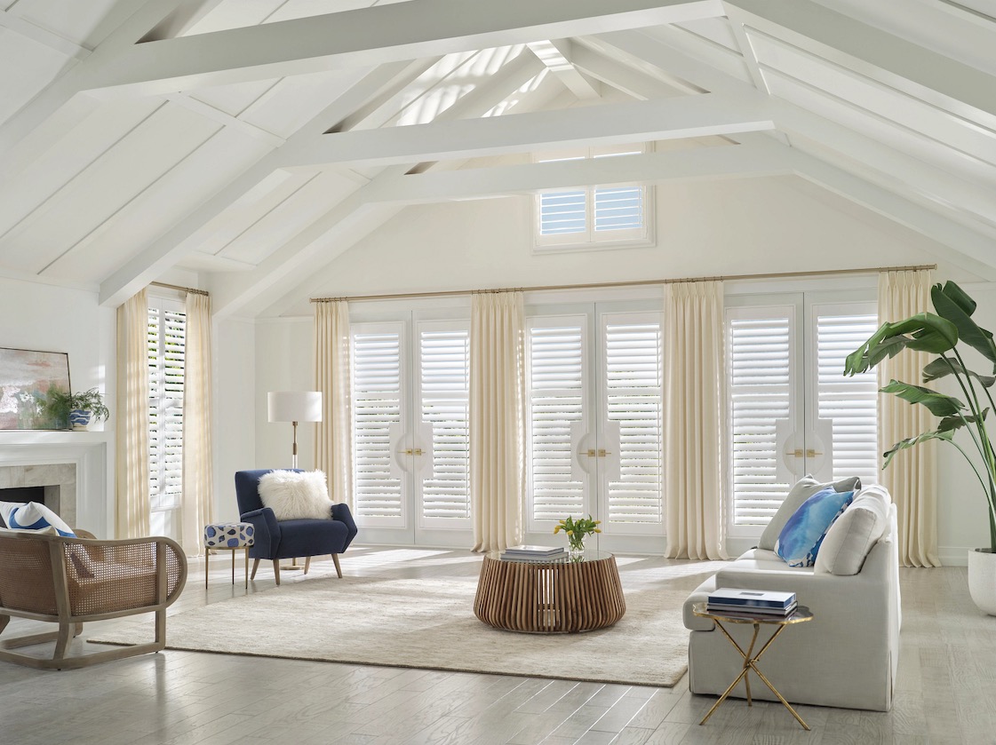 White plantation shutters on French doors and a transom window in a vaulted living room with cream drapery