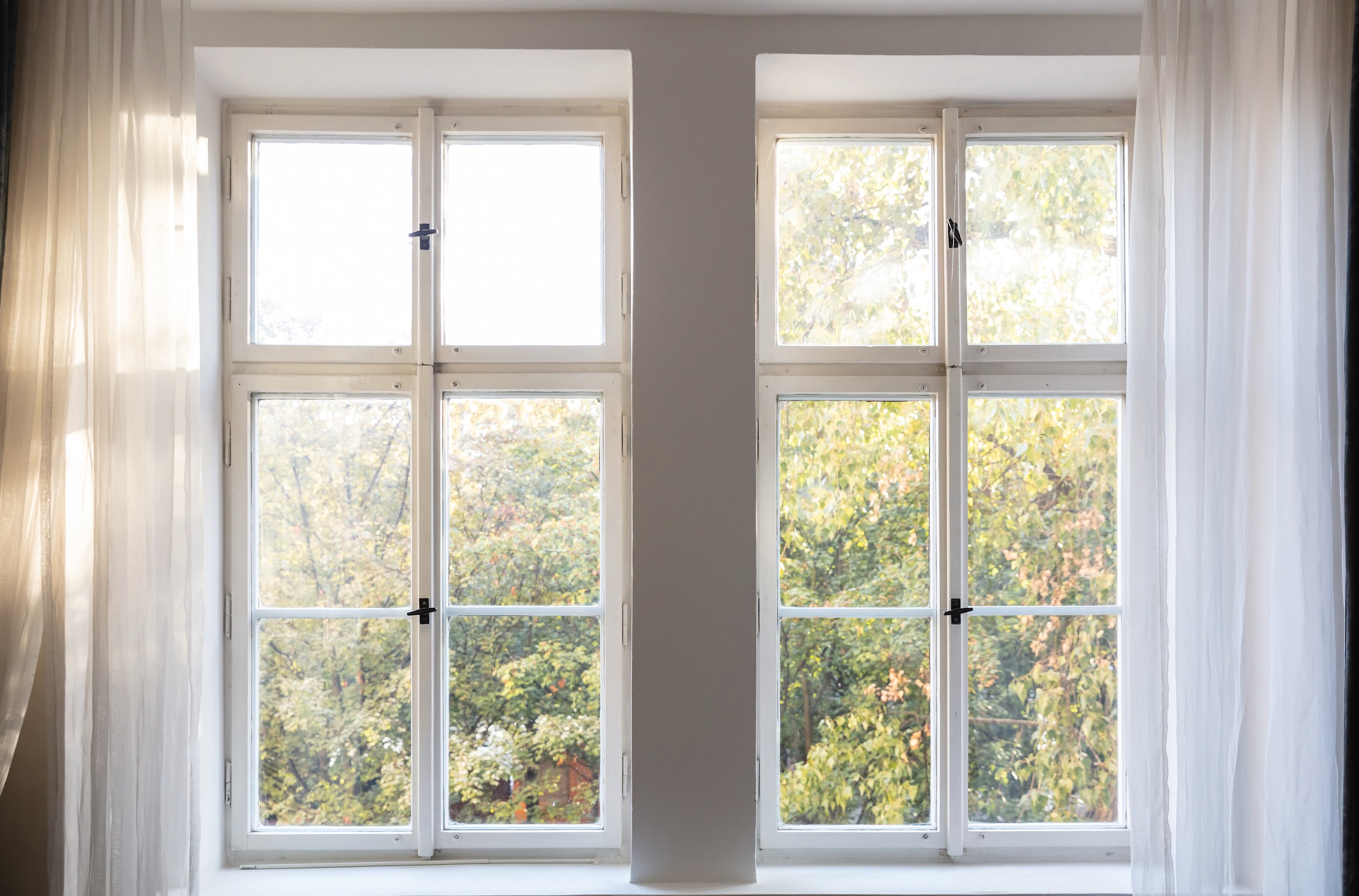 White sheer curtain panels framing bare double casement windows overlooking trees