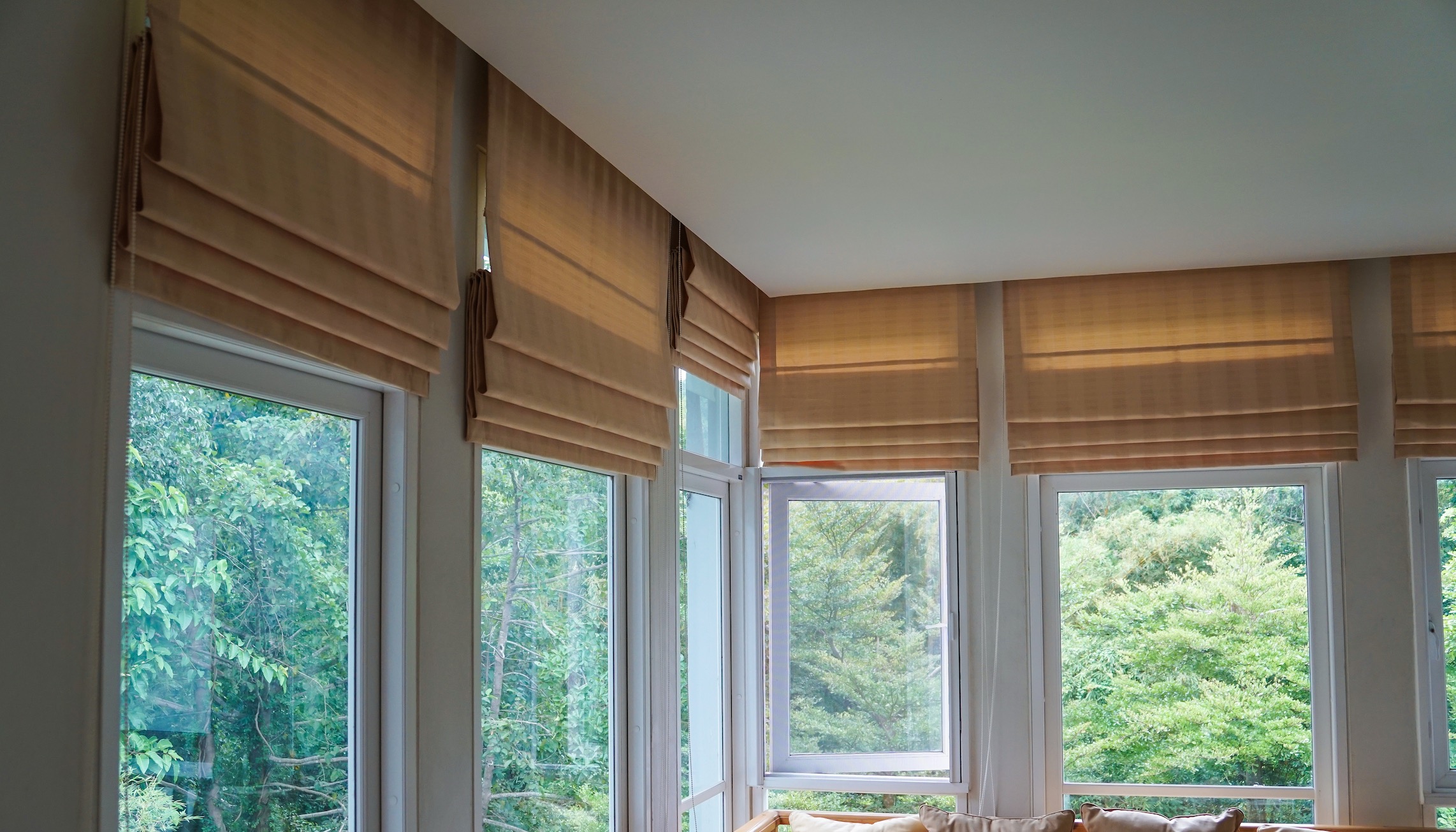Tan fabric Roman shades lowered across a corner bank of windows facing woods