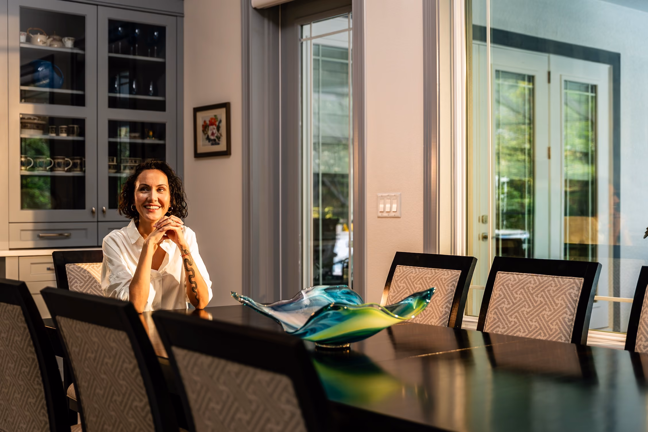 Smiling homeowner at a dark wood dining table beside glass patio doors and a glass art bowl