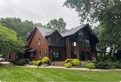 Large rustic wooden house with dark roof surrounded by green trees and shrubs under a cloudy sky.