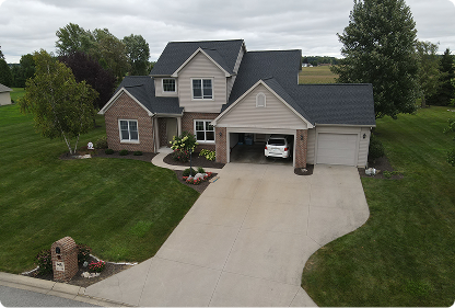 Two-story suburban house with attached garage and a white car parked inside, surrounded by green lawn and trees on a cloudy day.