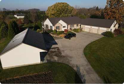 Aerial view of a suburban house with a large driveway, garage, and a black pickup truck parked near the entrance, surrounded by green grass and trees in autumn.