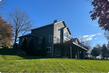 Side view of a two-story gray house with a large balcony, surrounded by green lawn and trees under a clear blue sky.