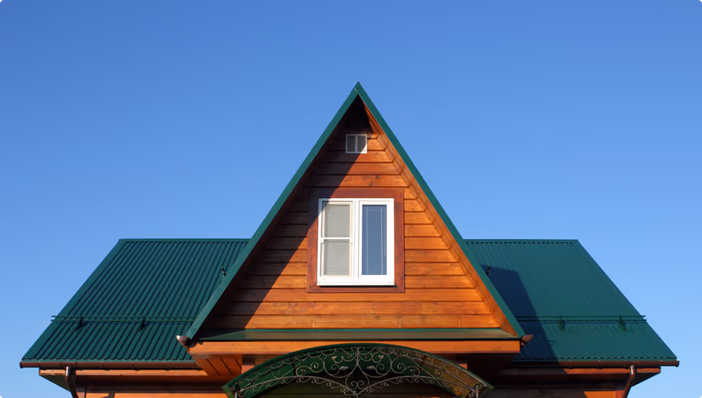 Front view of wooden house with green metal roof and triangular dormer window under clear blue sky.