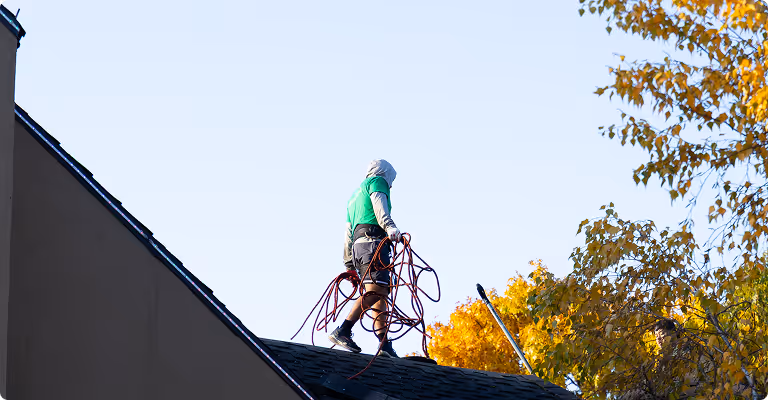 A person wearing a green shirt and hood carrying coiled red ropes walking on a sloped rooftop with autumn trees in the background.