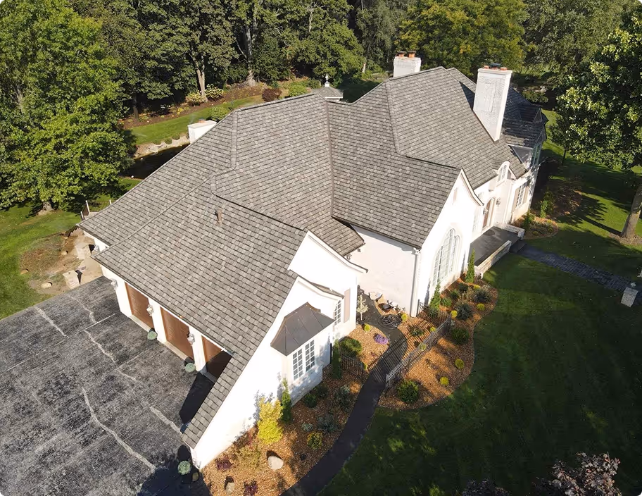 Aerial view of a large house with a gray shingled roof surrounded by green lawn and trees.