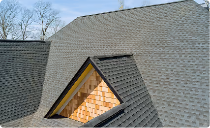 Close-up of a roof with dark gray architectural shingles and a triangular dormer with wooden siding inside the opening.