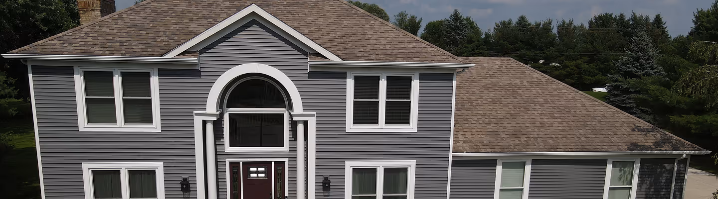 Two-story house with gray horizontal siding, white trim, and a brown shingled roof under a partly cloudy sky.