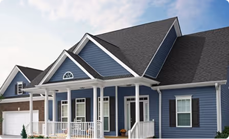 Blue house with dark roof, white porch columns, and vinyl siding under a partly cloudy sky.