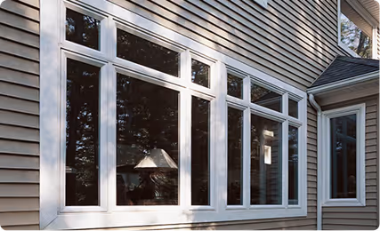 White-framed picture windows on a beige-sided house reflecting outdoor trees.
