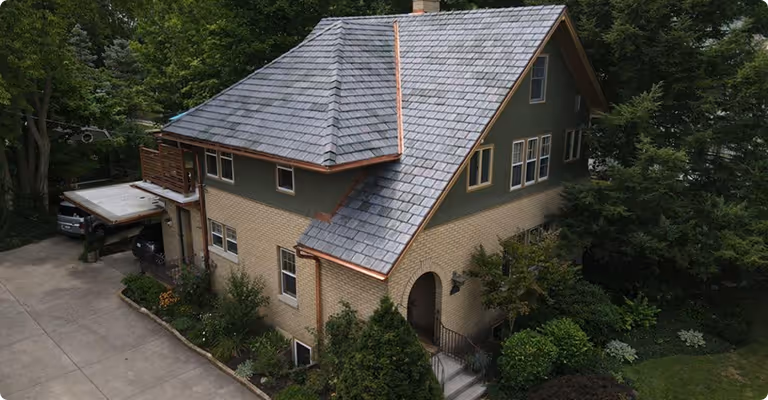 A two-story house with a gray shingle roof, beige brick walls, green siding, surrounded by trees and garden shrubs, with a driveway on the left.