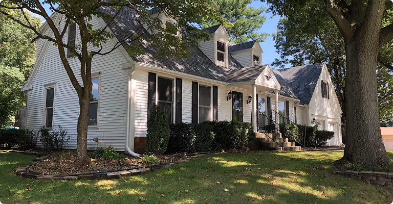 White two-story house with black shutters, dormer windows, and a front porch surrounded by a green lawn and trees.