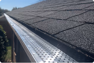 Close-up of a roof with asphalt shingles and a metal gutter guard installed along the edge.