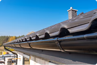 Close-up of a dark brown roof gutter system installed along a house roofline under clear blue sky.