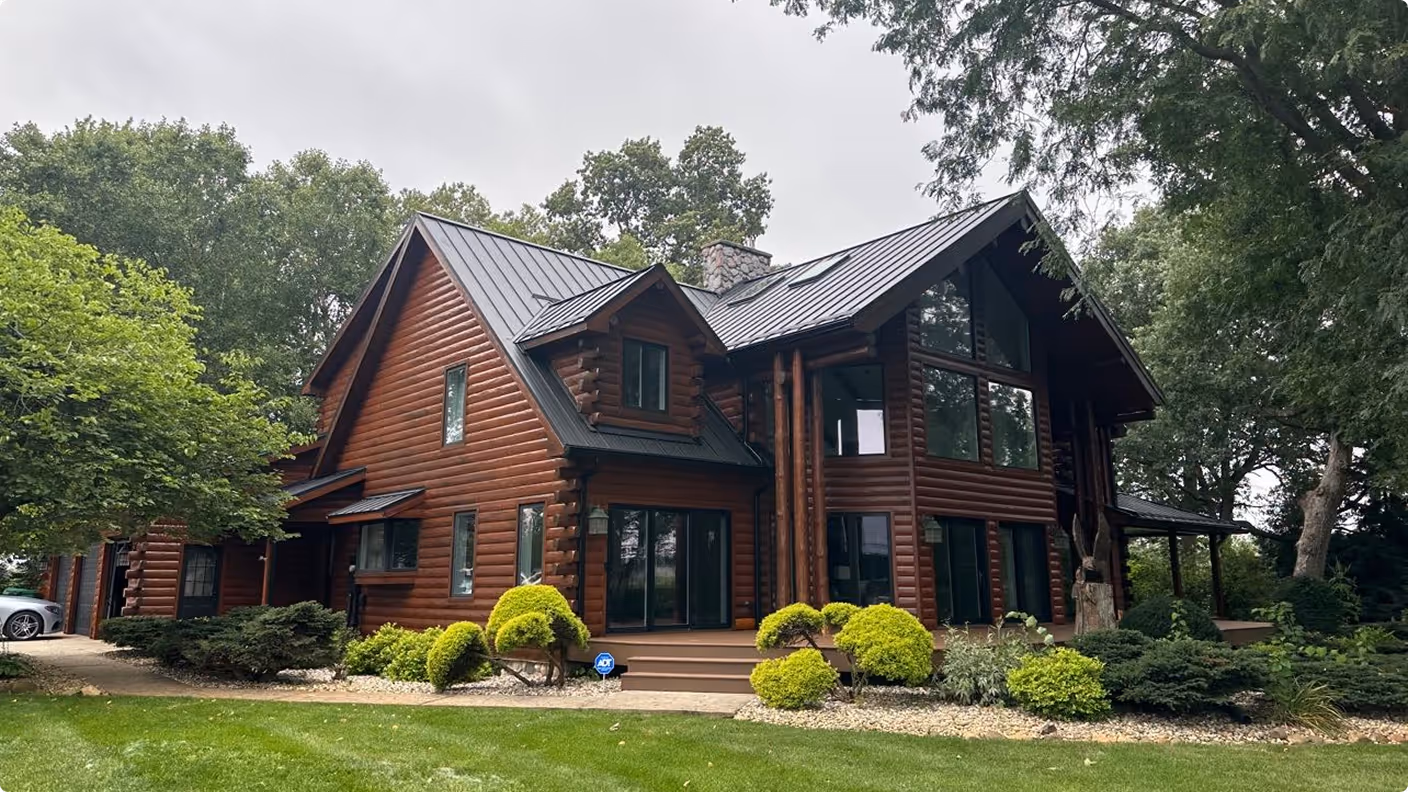 Two-story log cabin house with large windows, metal roof, and landscaped yard surrounded by trees.