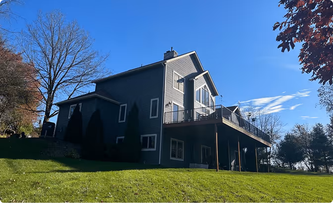 Two-story dark grey house with large windows and a raised wooden deck overlooking a green lawn under a clear blue sky.
