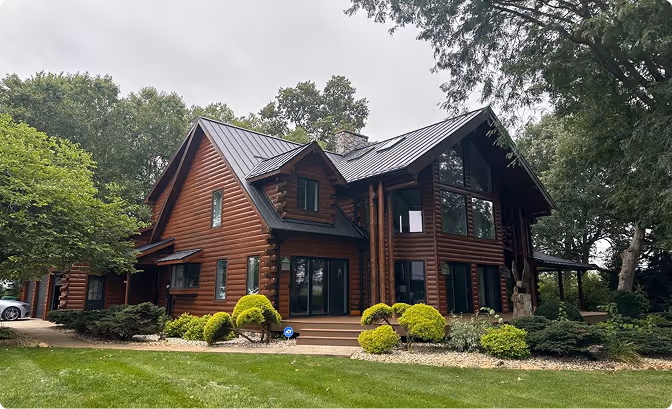 Large modern log cabin-style house with dark metal roof, surrounded by green trees and manicured lawn under an overcast sky.