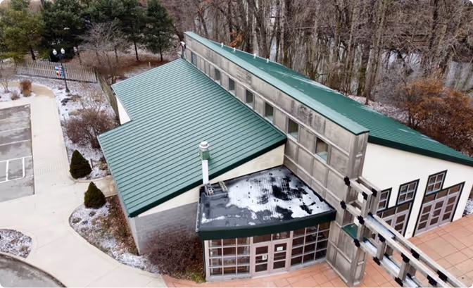 Aerial view of a modern building with green metal roofs, surrounded by a paved walkway and trees.