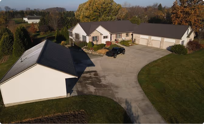 Aerial view of a large suburban house with a three-car garage, black SUV parked on the driveway, detached white garage, and surrounding green lawn and trees.