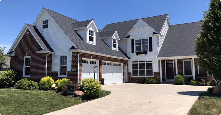 Large two-story suburban house with brick and white siding, two-car garage, and neatly landscaped front yard under a clear blue sky.
