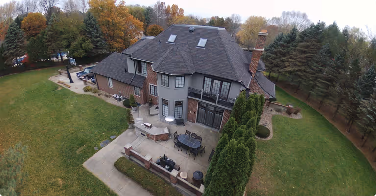 Aerial view of a spacious two-story brick house with a patio, outdoor dining set, barbecue grill, and surrounding green lawn with trees.
