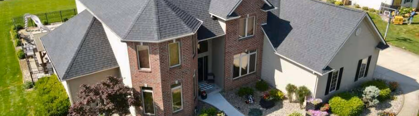 A large two-story suburban house with gray roof, brick facade, landscaped front yard, and a concrete driveway.