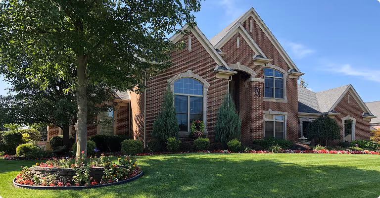 Large brick house with tall windows, landscaped front yard with green grass, flower beds, and a tree in a circular stone planter.