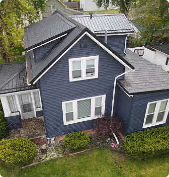 Two-story house with dark blue siding, white trim, multiple windows, and a gray metal roof seen from an elevated angle surrounded by green bushes and trees.