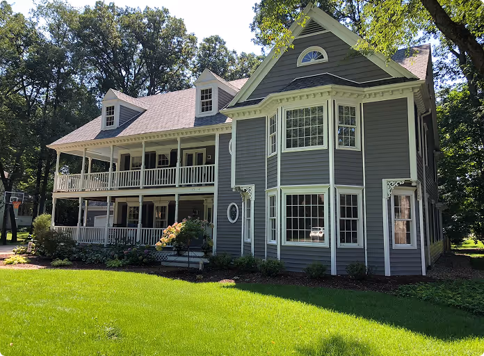 Large two-story gray house with white trim, bay windows, wrap-around porch, and green lawn surrounded by trees.