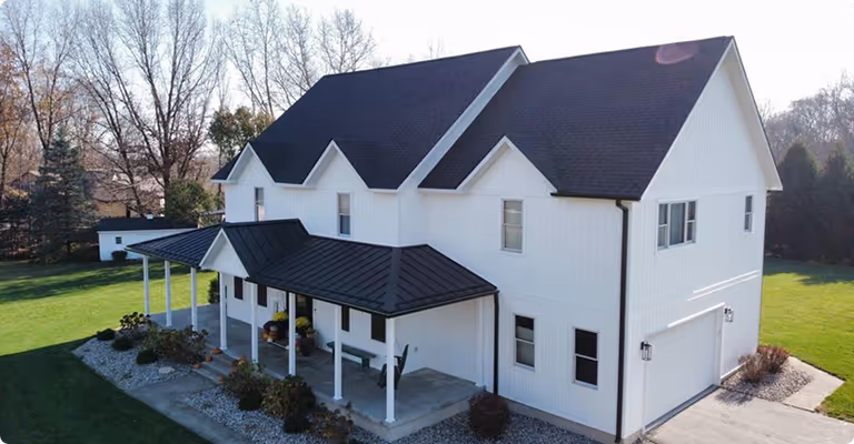 Two-story white house with black roof, front porch, and surrounding lawn and trees.