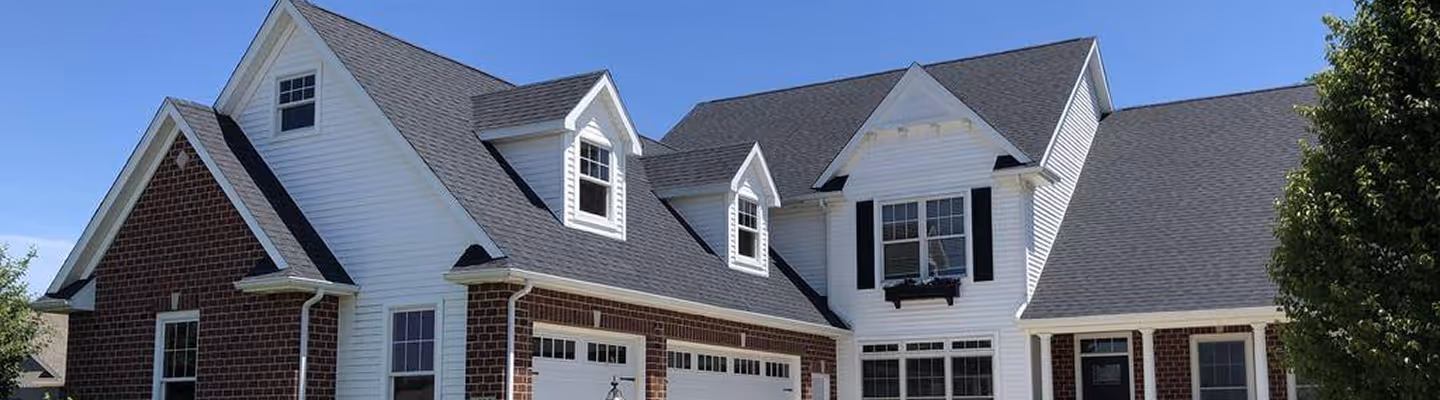 Large two-story suburban house with white siding, brick accents, dark shutters, and multiple dormer windows under a clear blue sky.